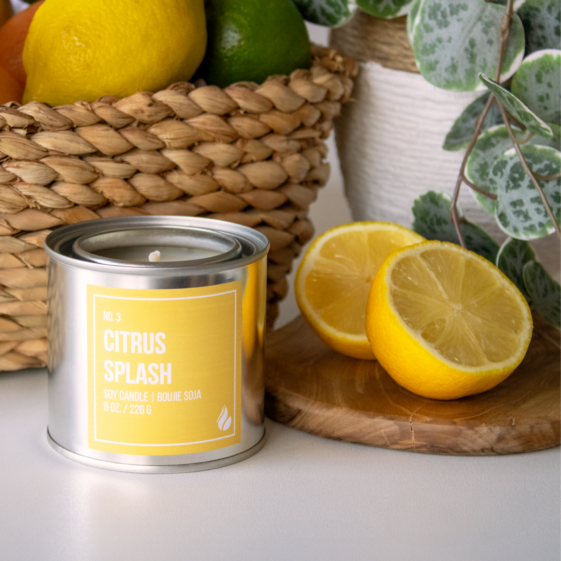 Paint can candle labeled 'Citrus Splash' next to citrus fruits and a woven basket on a white surface
