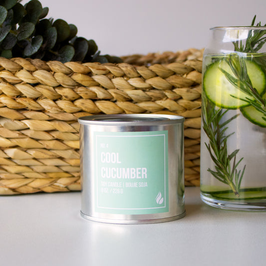 Product photograph of Cool Cucumber paint can candle with a basket of eucalyptus and cucumber water as props.