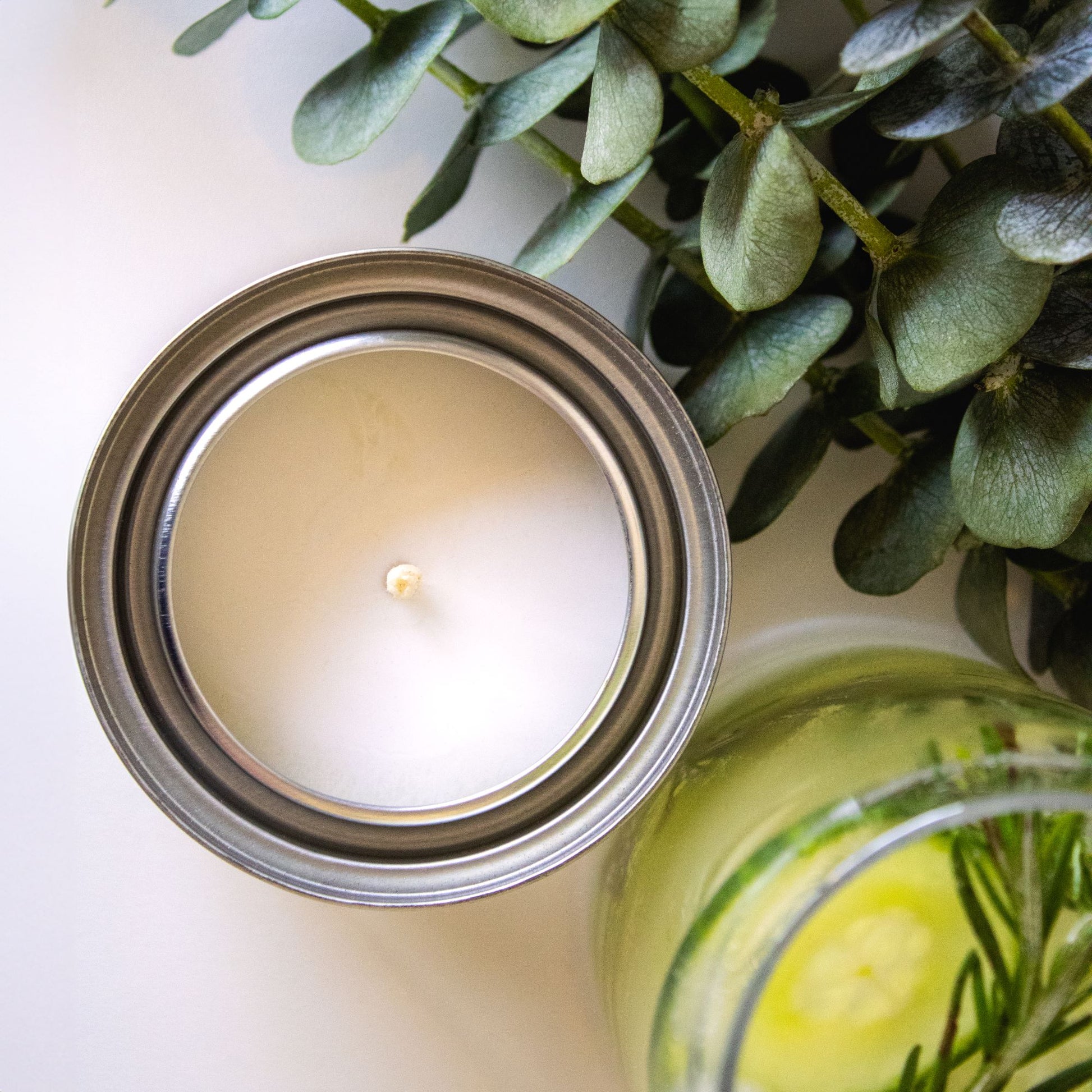 Cool Cucumber candle next to green leaves and cucumber water on a white table top.