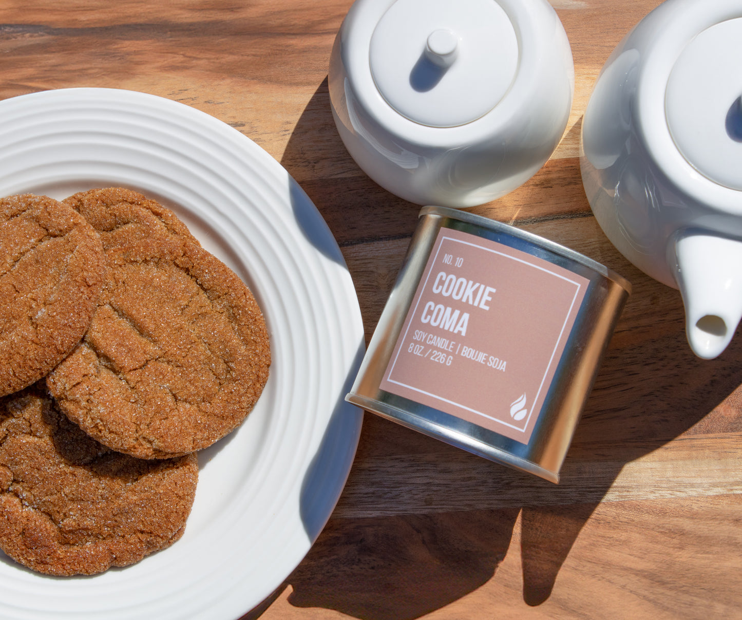 Product Photograph of Cookie Coma Paint Can Candle next to white dishware. White plate on the left features a plate of ginger cookies.