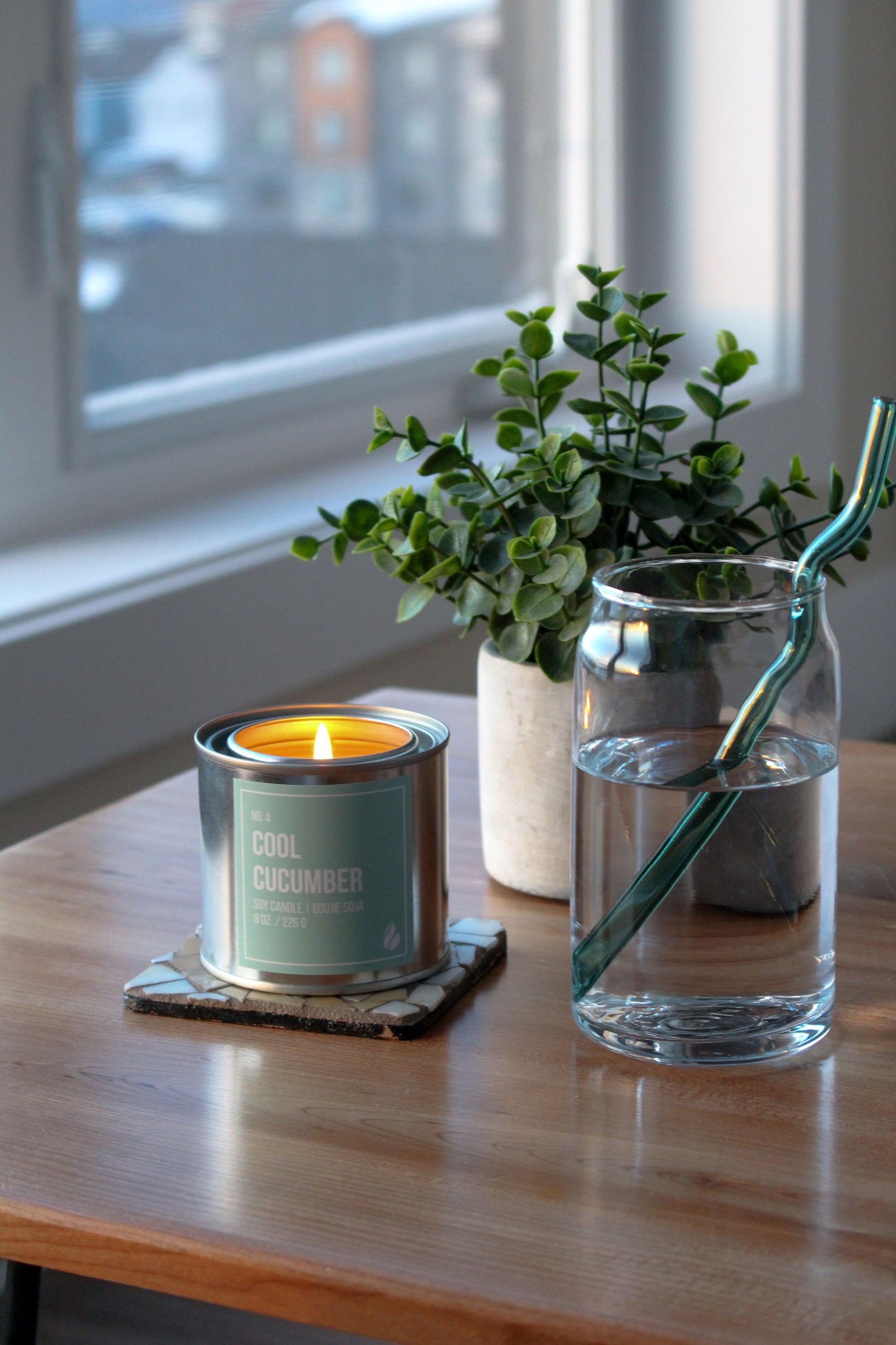 Cool cucumber paint can candle, plant, and glass with straw on a wooden surface near a window