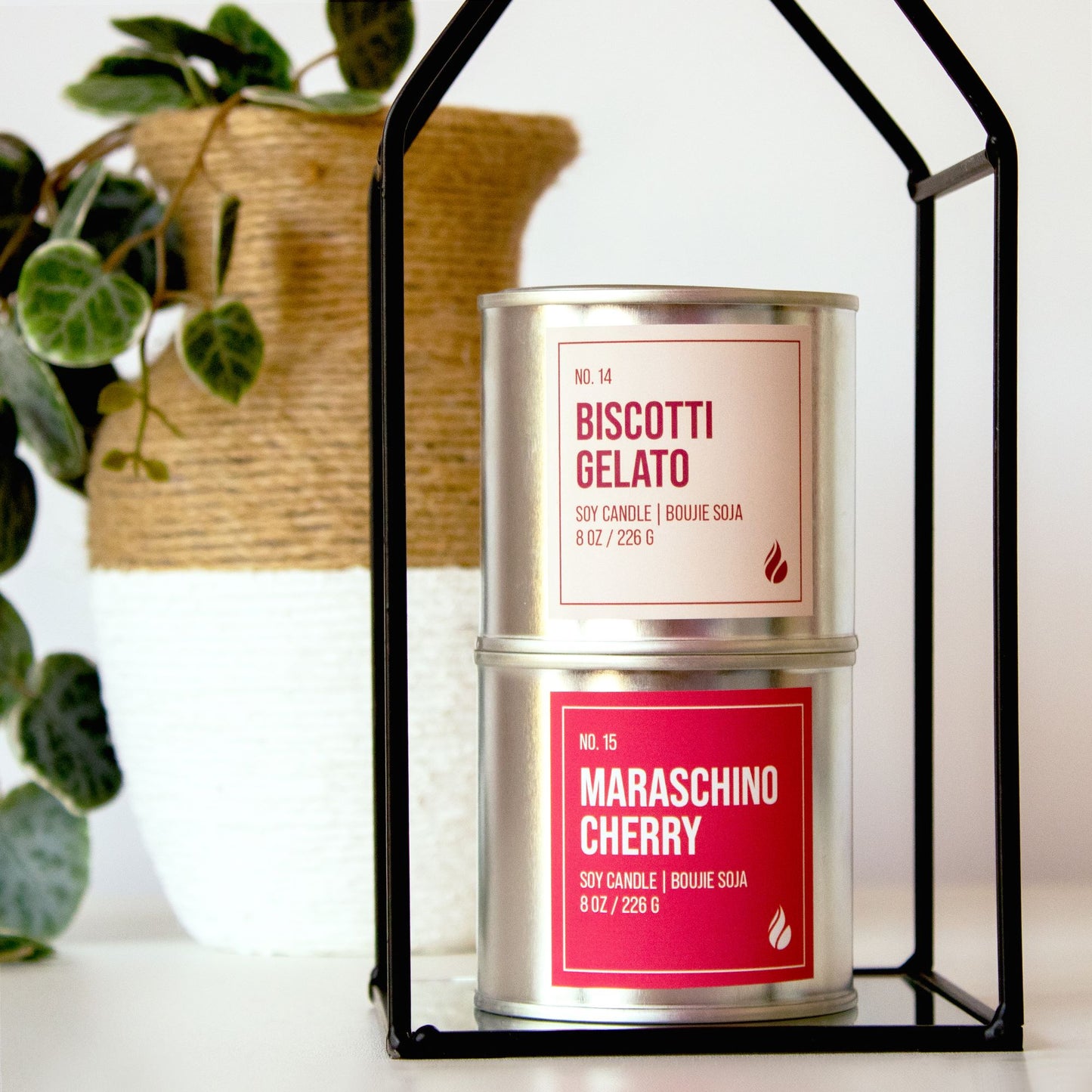 Photo of Biscotti Gelato candle placed on top of a Maraschino Cherry candle surrounded by props including a black wire sculpture in the shape of a house, and a plant in a brown and white colour block vase.
