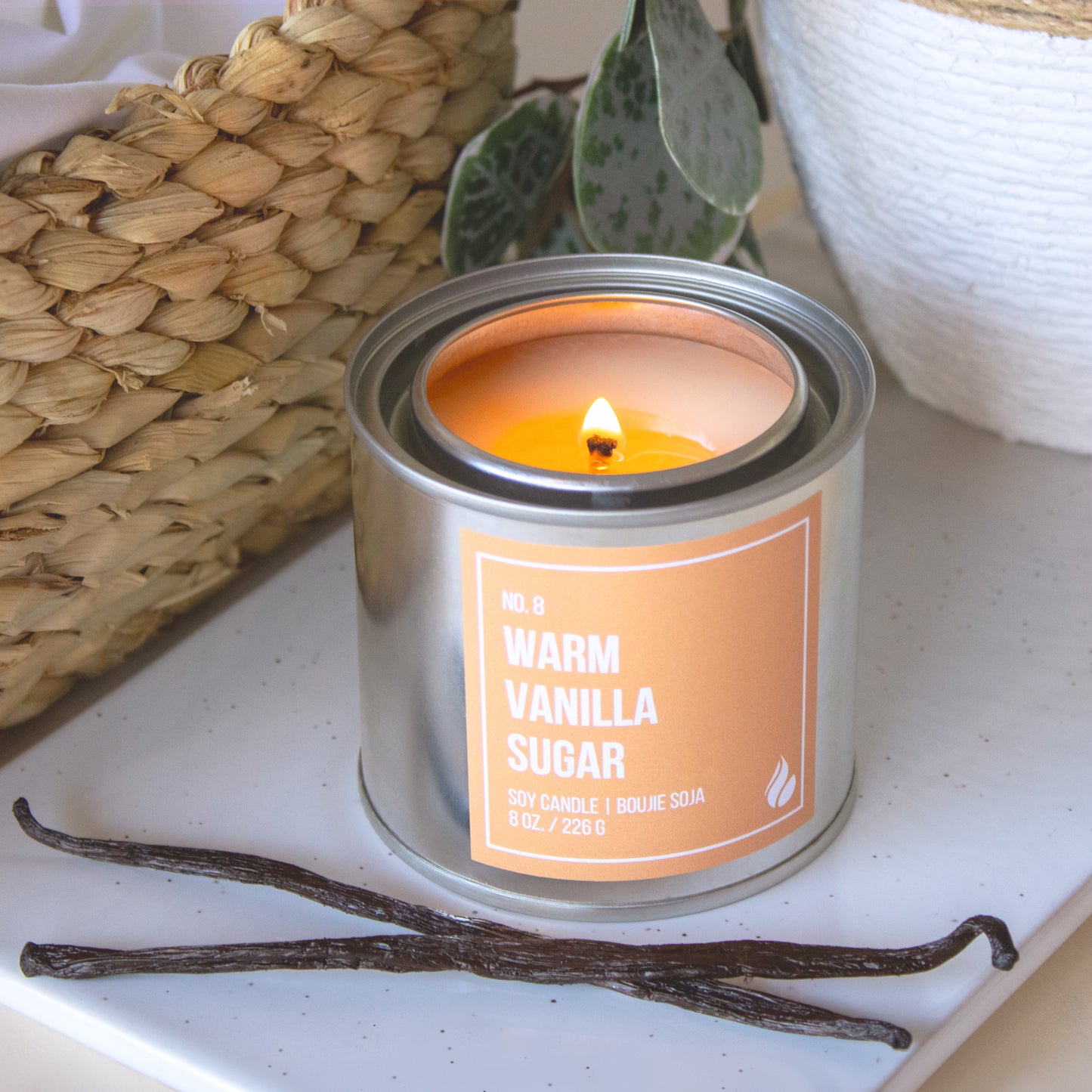 Product photograph of Warm Vanilla Sugar Paint Can Candle next to a basket, a white plant opt and two vanilla beans.