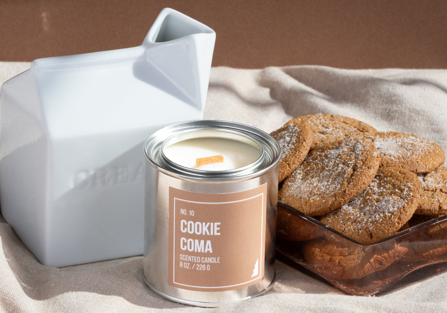 A picture of a Cookie Coma scented Paint Can Candle on a clothed table featuring brown walls, a cup of milk and a container of cookies..